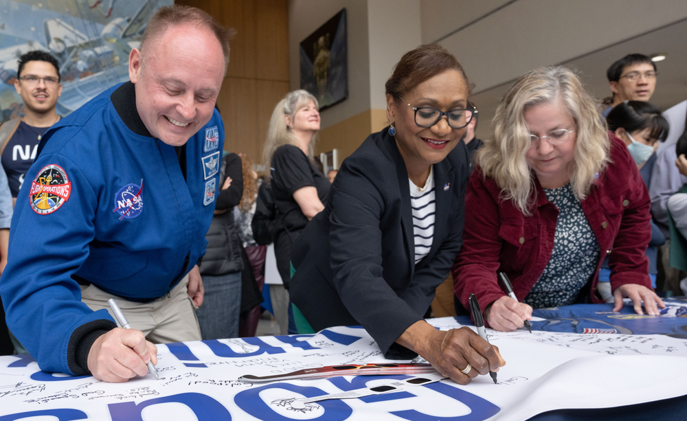 Three people sign a banner on a table in a busy lobby. 