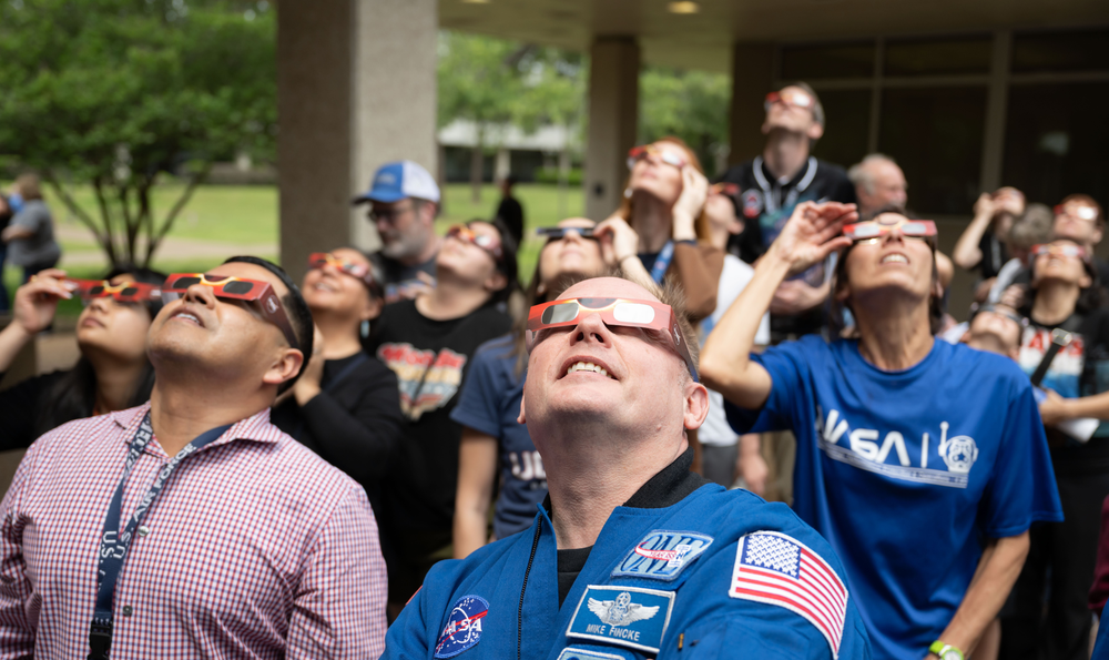 A crowd of people outside gaze at the sky wearing eclipse glasses. A man wearing a blue flight suit stands in the front.