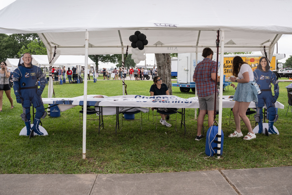 A person sitting at a booth outside with two people on her left signing a banner.