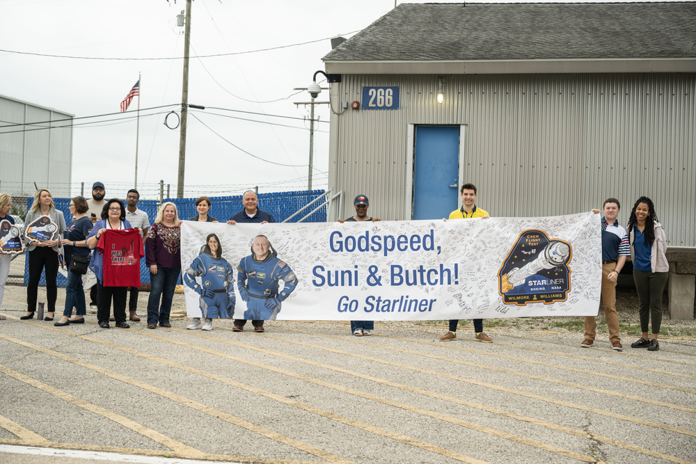 A group of people hold a large banner reading Godspeed, Butch and Suni while standing on the side of a road.