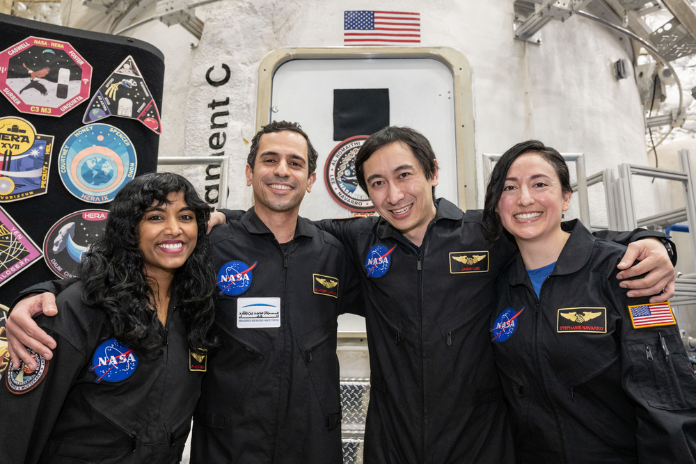 A group of four smiling NASA personnel in black jumpsuits stands together in front of a spacecraft module. Each individual has a NASA patch and name tag on their suit.