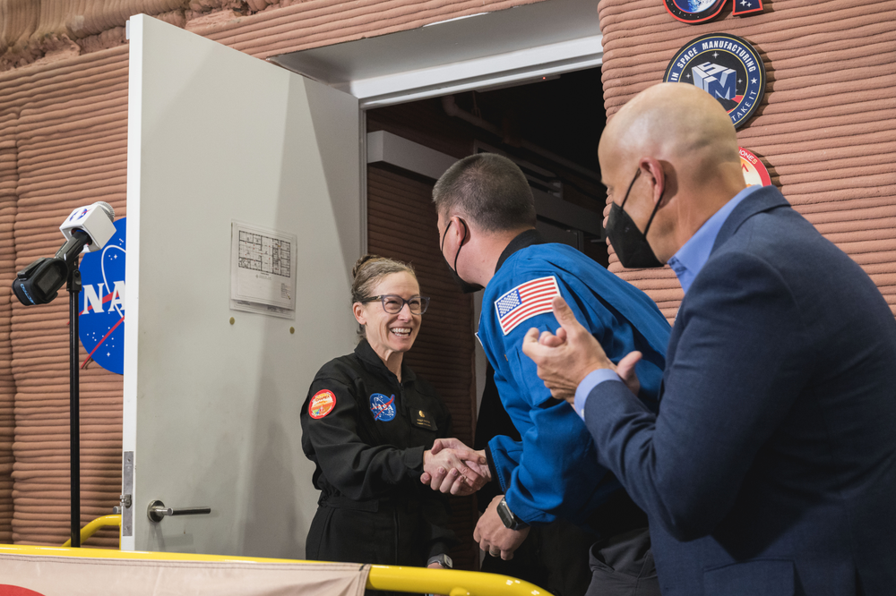 A woman in a black NASA jumpsuit is shaking hands with a man in a blue NASA astronaut suit. Another man in a blue suit is clapping. They are standing at the entrance of a building with NASA logos and other mission patches on the wall.