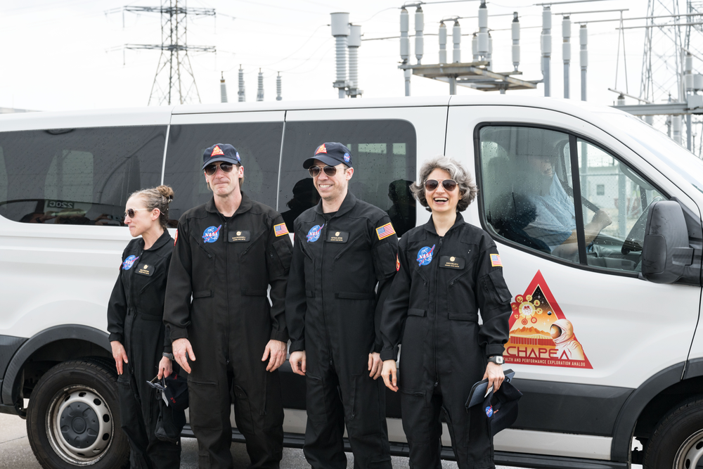 Four people wearing black NASA jumpsuits are standing in front of a white van with a CHAPEA mission logo. The individuals are wearing sunglasses and caps, and the background shows industrial equipment and power lines.