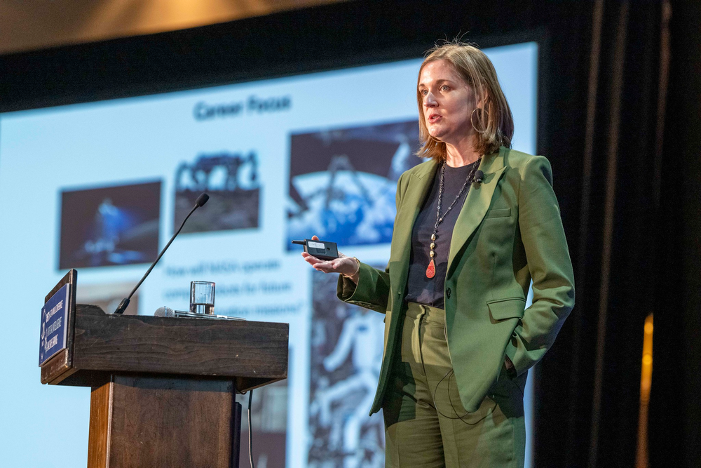 A person in a green suit gives a presentation on a stage with a screen behind her.