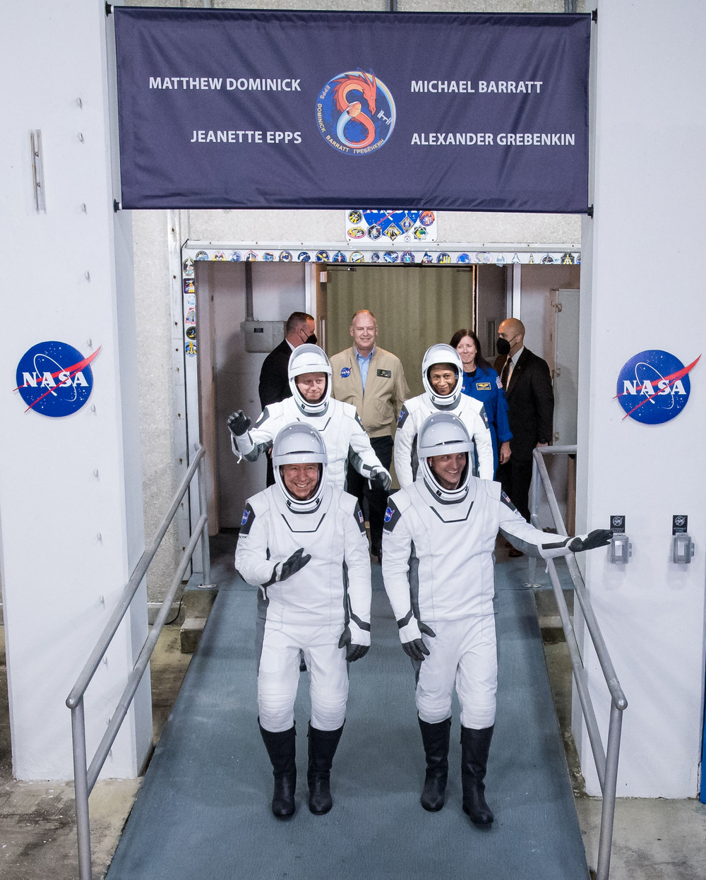 Four people in white flight suits wave and smile while walking out of a white building.