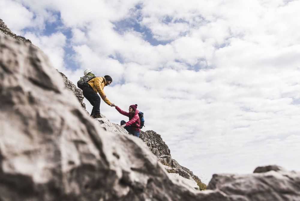 A man wearing a yellow jacket helps a woman wearing a pink jacket climb up a rocky mountainside.