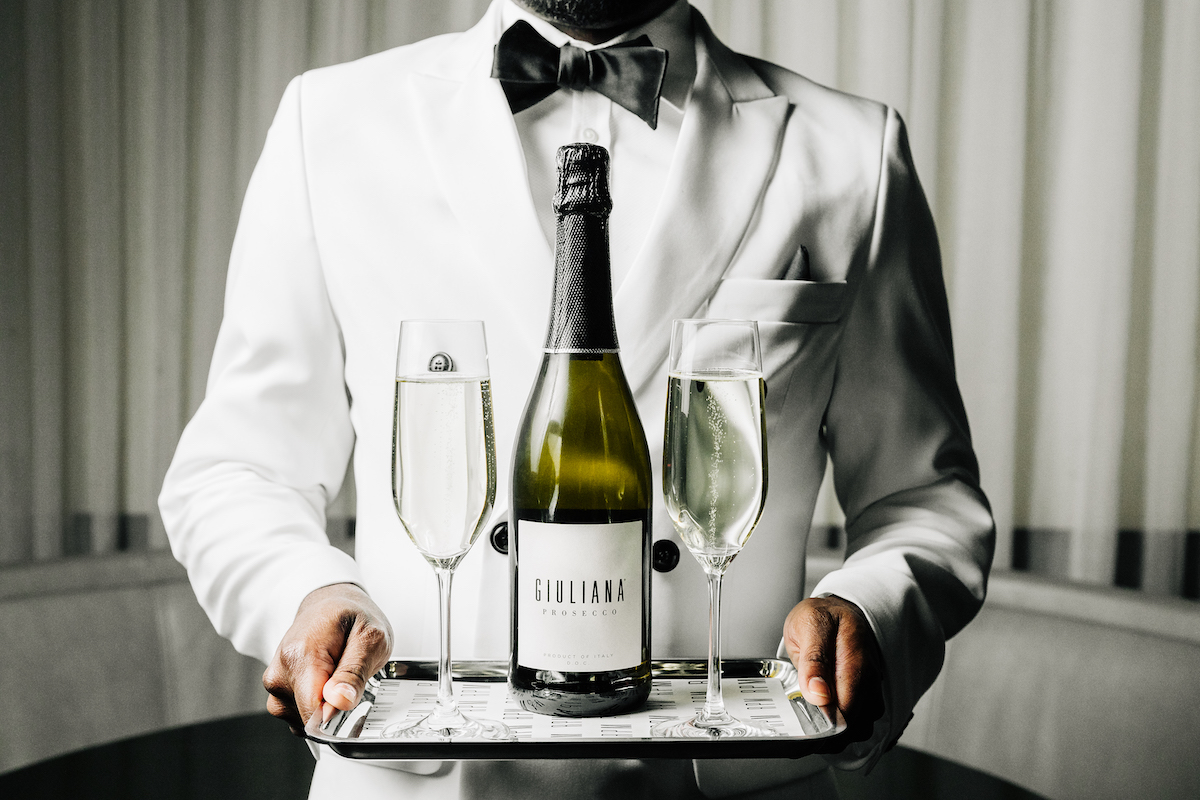 Server in white coat jacket with a tray of two champagne flutes and a bottle of champagne. 