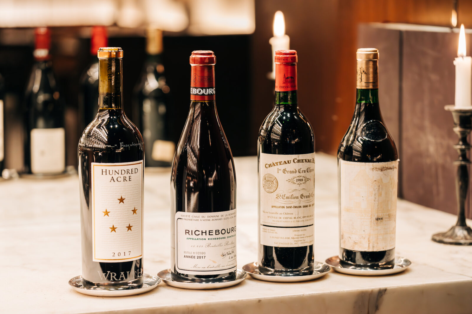 Four bottles of wine on a marble table. Wine labels include Hundred Acre, Richebourg, Château Cheval Blanc, and one with an unreadable label.