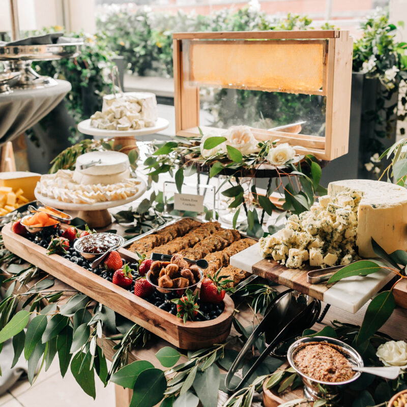 Assorted food display with cookies, cake, cheese, crackers and jams surrounded by greenery.