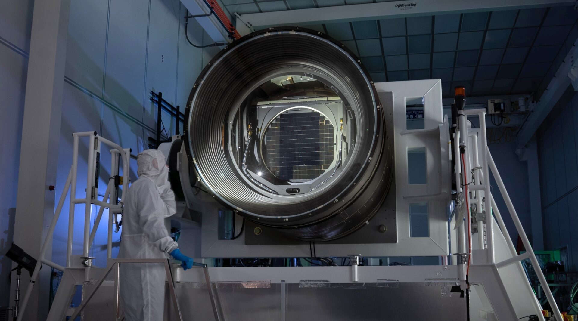 The LSST Camera, the largest camera ever built for astronomy. The camera opening is a large black ring with glass lenses, a bit smaller than a person is tall. In the center of the opening is the camera's focal plane detector, which is made of 189 square CCD chips arranged in a roughly square shape. The camera is suspended on a white metal frame with white handrails. A person in head to toe white clean room clothing shines a flashlight into the camera opening.