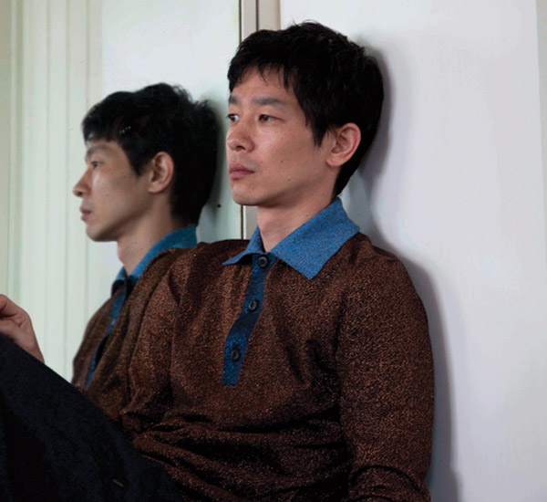 Ryo sits next to his reflection in the mirror. He is wearing a Lurex polo shirt by Prada