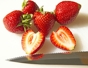 Image showing A sunlit sliced strawberry rests with other whole strawberries with a knife on a white cutting board (macro-14MP camera)
