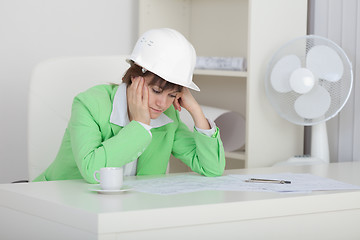 Image showing Sad woman engineer with helmet on head sits at table on workplac