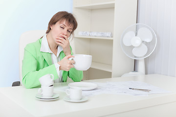 Image showing Tired woman sits at table with big cup of coffee and yawns