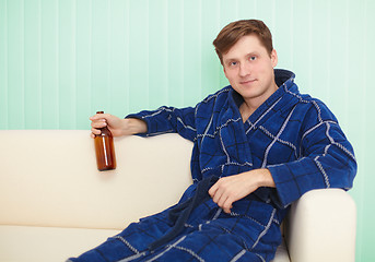 Image showing Young man drinks beer in dressing gown on sofa