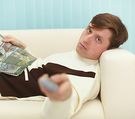 Image showing Young man, lying on couch with magazine and TV remote control