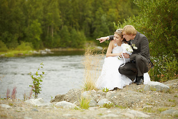 Image showing Bride and groom sit on riverbank