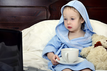 Image showing little girl in a bathrobe relaxing on the bed after a shower with cup of tea and laptop