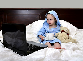 Image showing little girl in a bathrobe relaxing on the bed after a shower with cup of tea and laptop