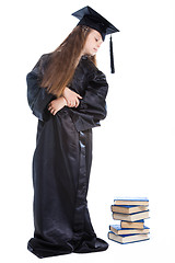Image showing girl in black academic cap and gown looking at the pile of books