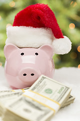 Image showing Pink Piggy Bank Wearing Santa Hat Near Stacks of Money on Snowfl