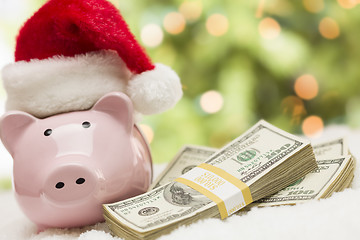 Image showing Pink Piggy Bank Wearing Santa Hat Near Stacks of Money on Snowfl