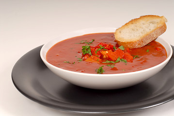 Image showing Tomato, red pepper, basil soup in white bowl with bread on a lig