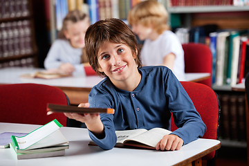 Image showing Cute Schoolboy Giving Book While Sitting At Table In Library