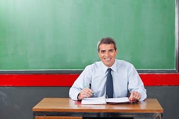 Image showing Confident Male Teacher With Pen And Binder Sitting At Desk