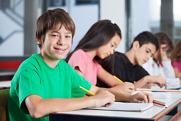 Image showing Boy Sitting At Desk With Friends Writing In Classroom