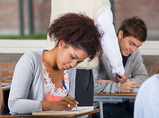 Image showing Student Writing Exam While Teacher Explaining Classmate In Backg