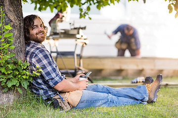 Image showing Carpenter Holding Mobilephone And Cup While Leaning On Tree Trun