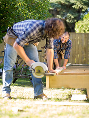 Image showing Carpenter Assisting Coworker In Cutting Wood With Handheld Saw