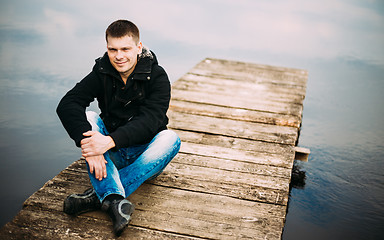 Image showing Young handsome man sitting on wooden pier, relaxing,  thinking. 