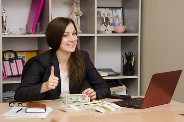 Image showing Pretty teenage girl sitting at desk with a pile of money and shows thumb