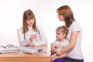 Image showing A pediatrician looks at the thermometer, sitting next to a woman with a child on her lap