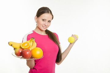 Image showing Girl athlete holding a plate of fruit and dumbbell