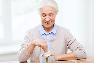 Image showing senior woman putting money into glass jar at home