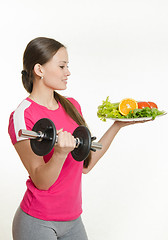 Image showing Sportswoman looking at a plate of fruit while holding a dumbbell in the other hand