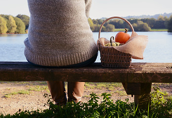 Image showing Woman sits on bench with a basket of pumpkins by a lake