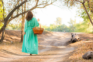 Image showing A girl in a long dress holding a basket is on the road into the distance