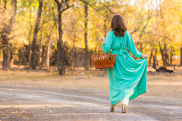 Image showing A girl in a long dress walking along the road with a basket in the hands of