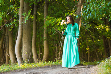 Image showing A girl in a long dress walking along the road and sniffing a bouquet of roses