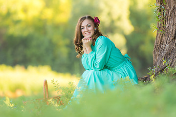 Image showing A girl in a long dress smiling while sitting against a tree in the woods