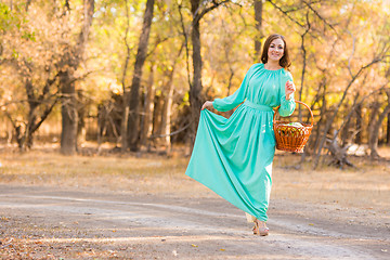 Image showing A girl in a long dress holding her dress and a basket of walking on the road