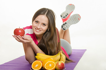 Image showing Sportswoman lying on a mat, holding an apple in hand, it is faced with a plate of fruit