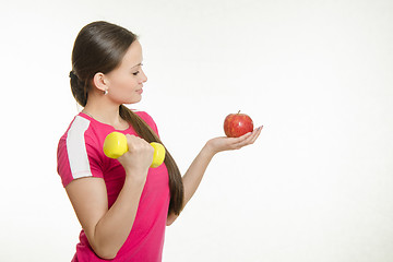 Image showing Athlete shakes muscles of the right hand dumbbell and looking at an apple in her left hand