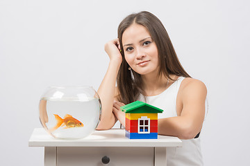 Image showing The girl sits at a table on which there is an aquarium with goldfish and toy house