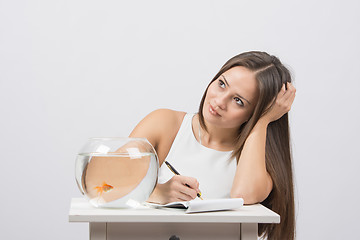 Image showing Girl thoughtfully writing in a notebook, standing next to an aquarium with goldfish