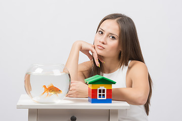 Image showing Thoughtful girl sitting at a table on which there is an aquarium with goldfish and toy house